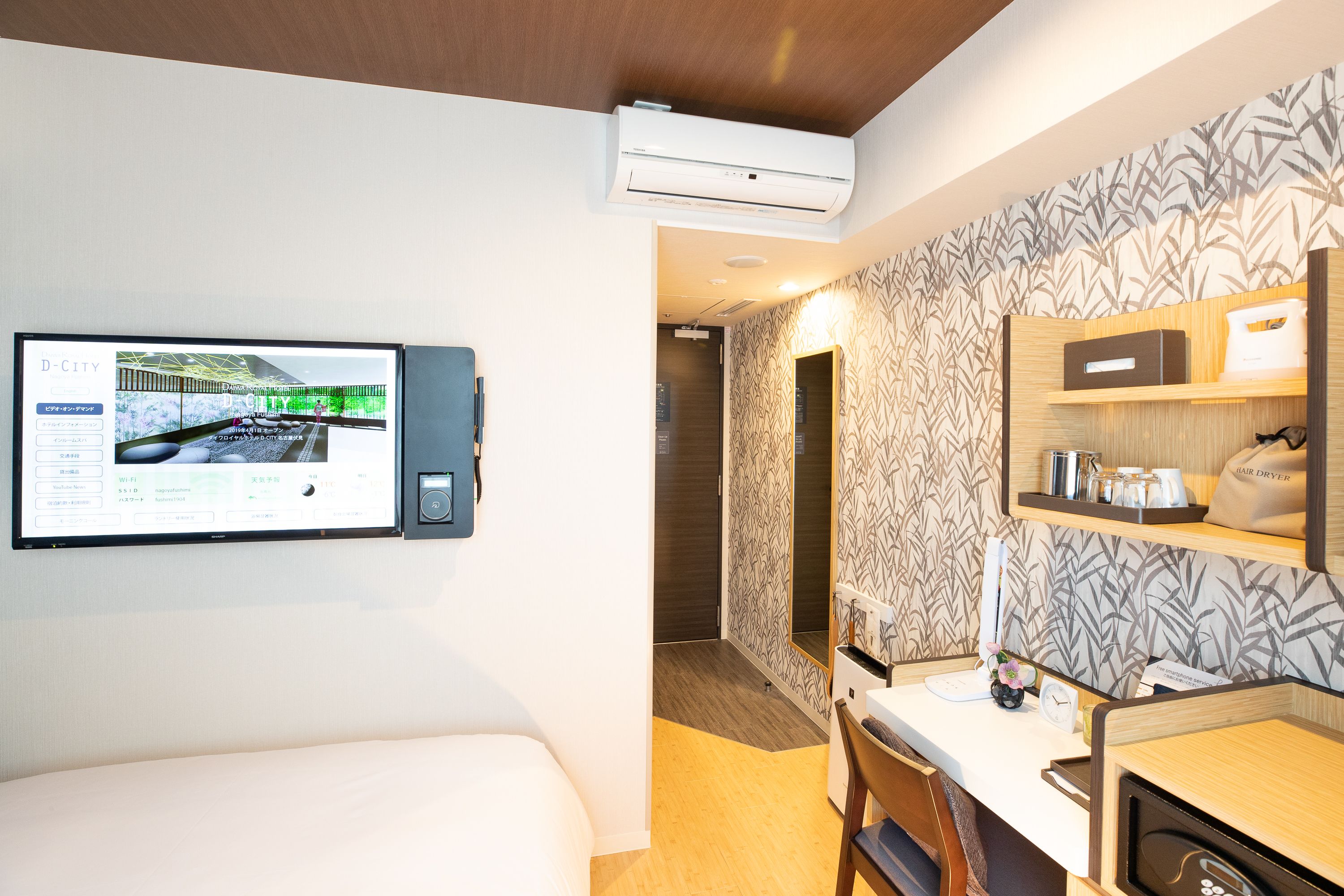 A hotel room with a bed, a desk, a mirror, and a wall-mounted TV displaying a cityscape.