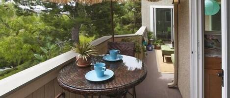 Un balcon avec une table en osier, des tasses bleues, une plante en pot et un parasol à toiture de chaume.
