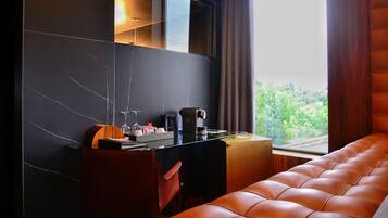 A modern hotel room with a dark marble wall, a wooden desk, a leather sofa, and a large window with a view of greenery.