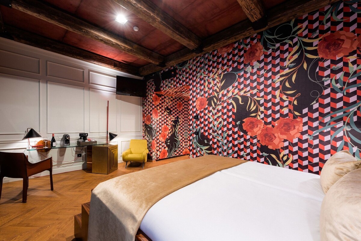 Una habitación de hotel con una cama, un escritorio con silla, una mesita con lámpara y una pared con estampado floral geométrico.