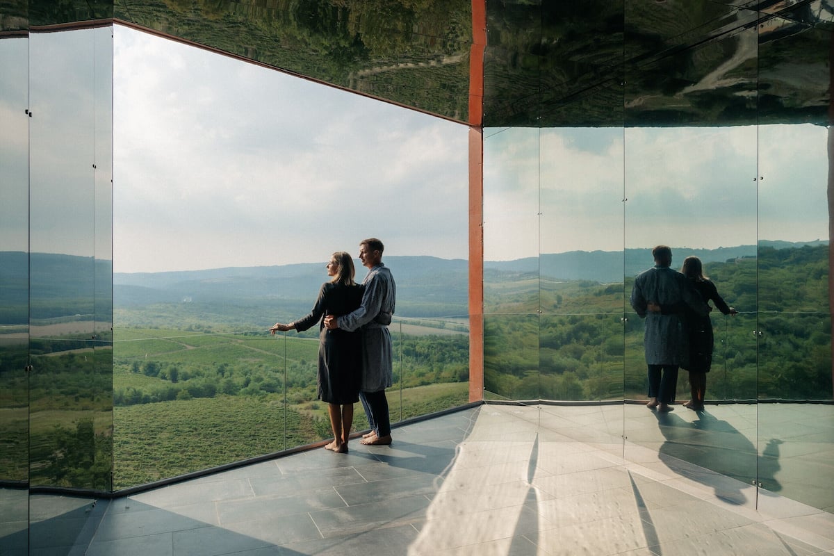 A couple standing in a room with large glass walls, overlooking a green landscape.