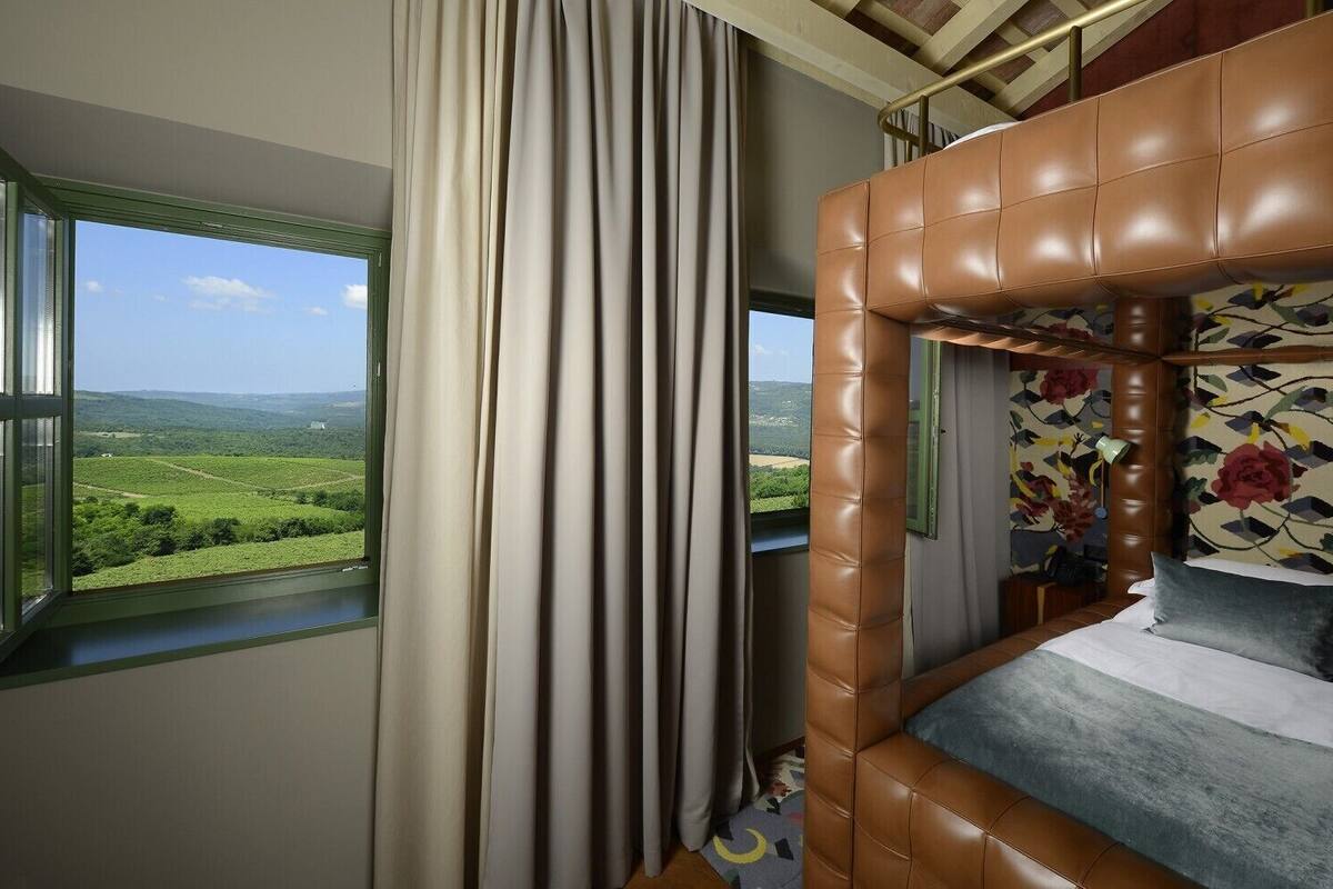 A bedroom with a view of a green landscape, a window with curtains, and a brown headboard.