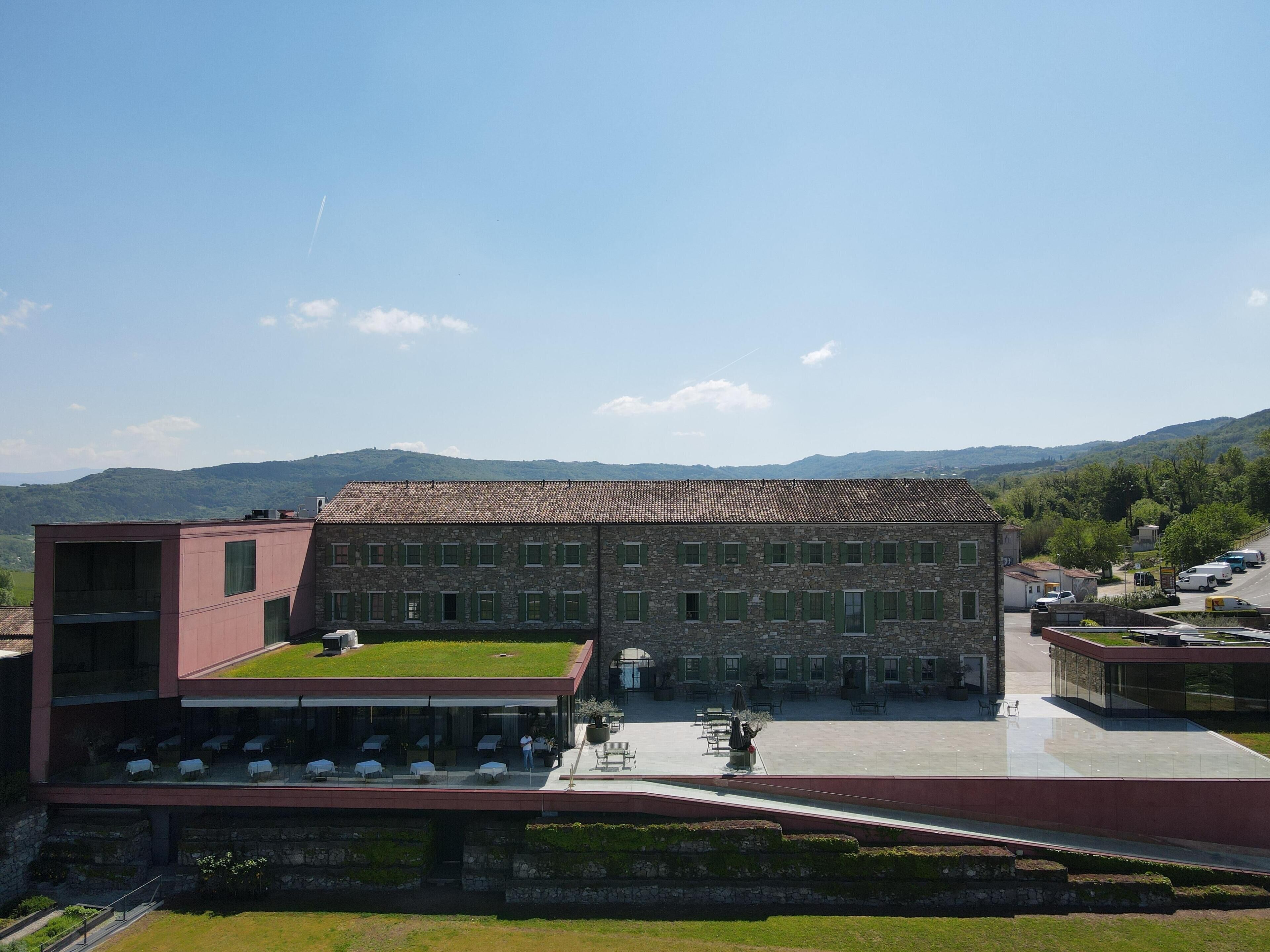 Un edificio con techo verde, un patio con mesas al aire libre y vistas a colinas al fondo.