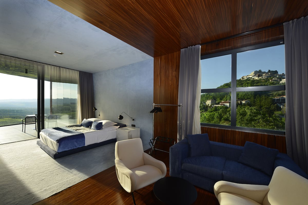 A modern hotel room with a large bed, a sofa, and a view of a hill with buildings.