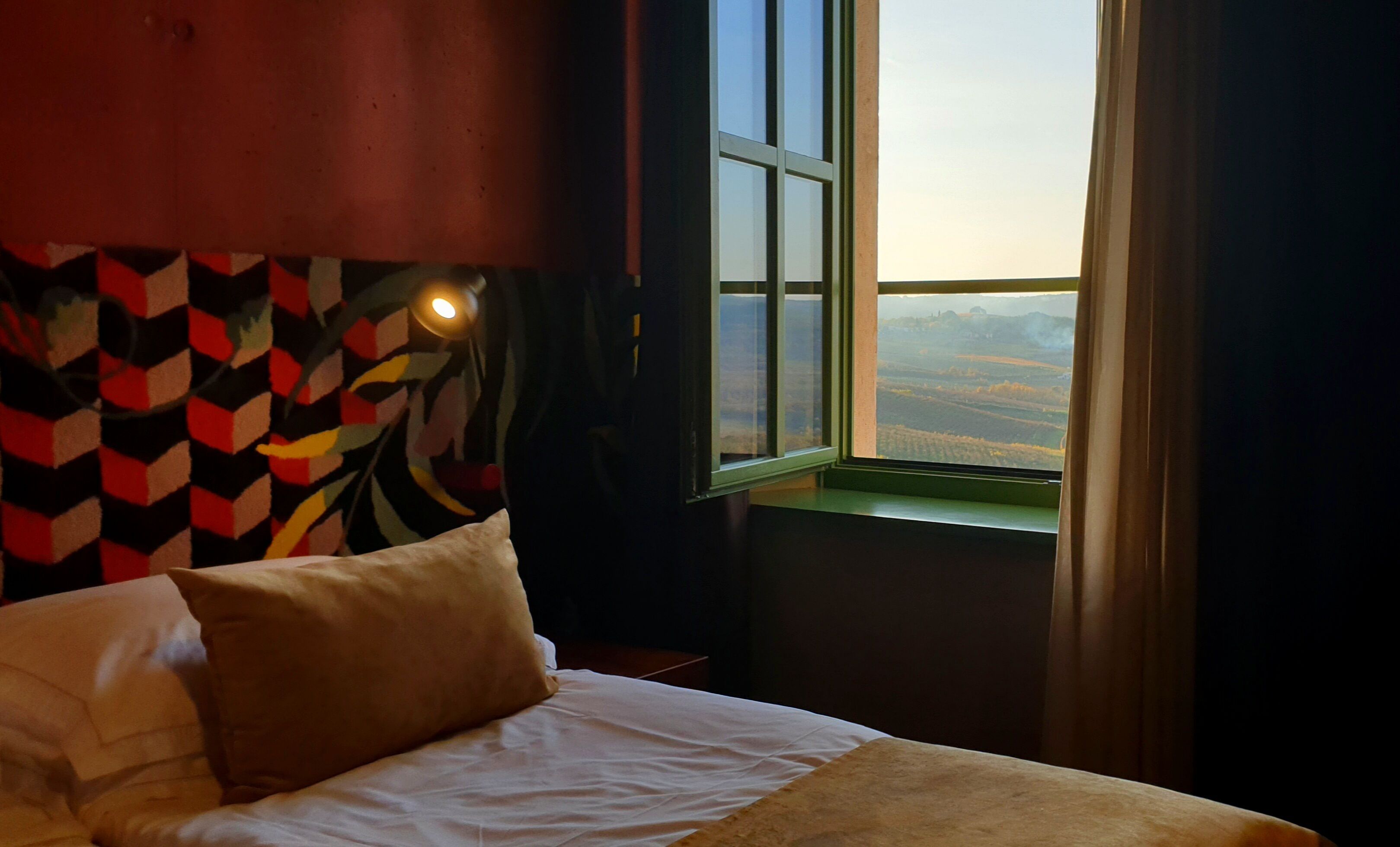 Una camera d'albergo con un letto, una finestra vista campagna e una parete con un motivo geometrico colorato.