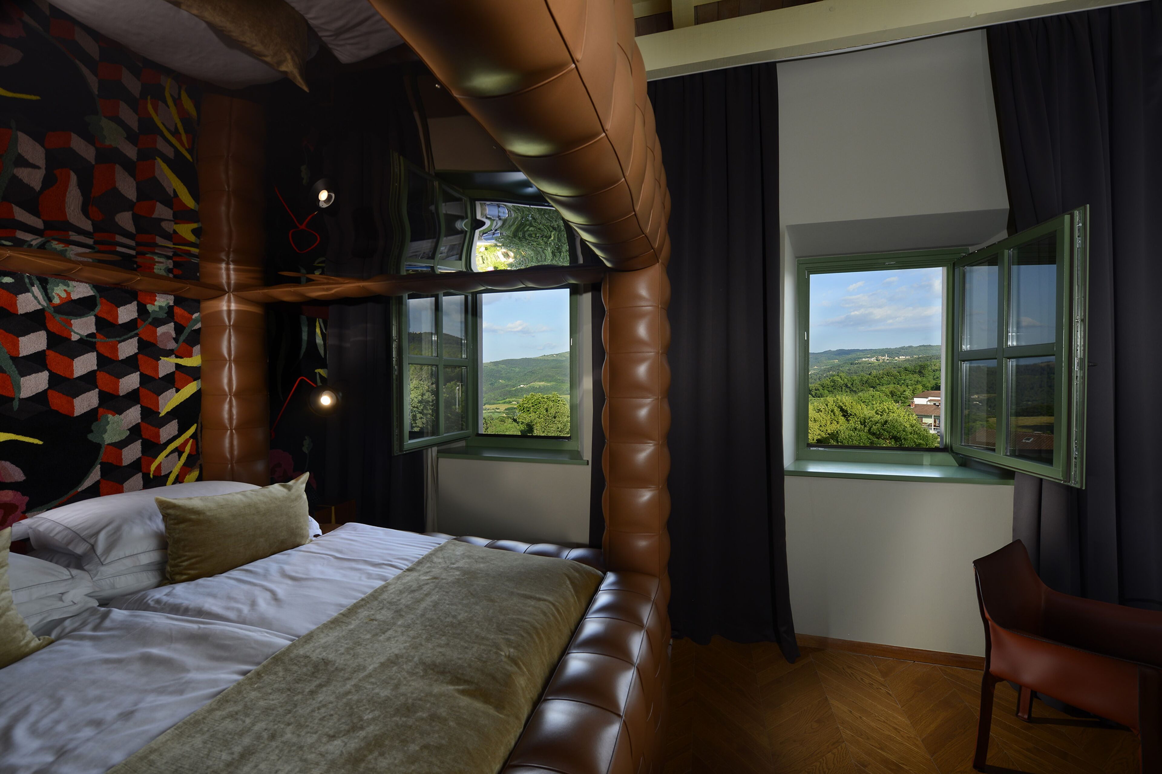 Ein Schlafzimmer mit einem großen Bett, zwei Fenstern, Blick auf Grünflächen und einem einzigartigen, geometrischen Wanddesign.