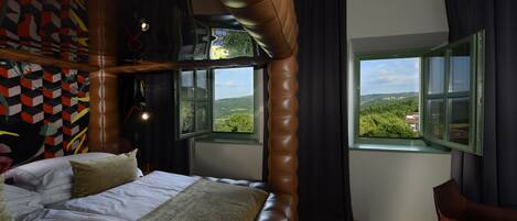 Ein Schlafzimmer mit einem großen Bett, zwei Fenstern, Blick auf Grünflächen und einem einzigartigen, geometrischen Wanddesign.