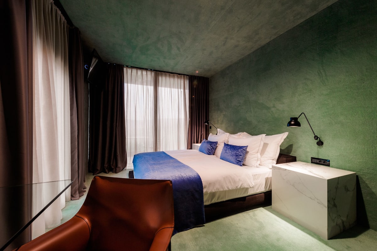 A modern hotel room with a large bed, a nightstand, a desk, and a leather chair.