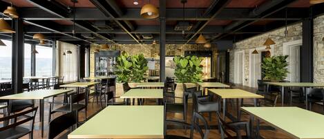 A modern restaurant with high ceilings, exposed beams, and large windows offering views. Tables are arranged in a spacious layout with chairs.