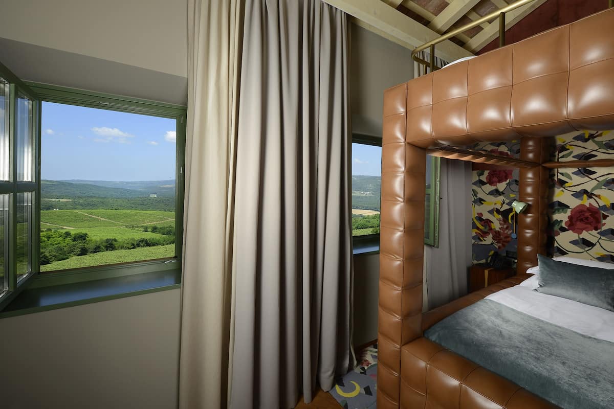 A room with a large window offering a view of a green landscape, a brown leather headboard, and a floral-patterned wall.