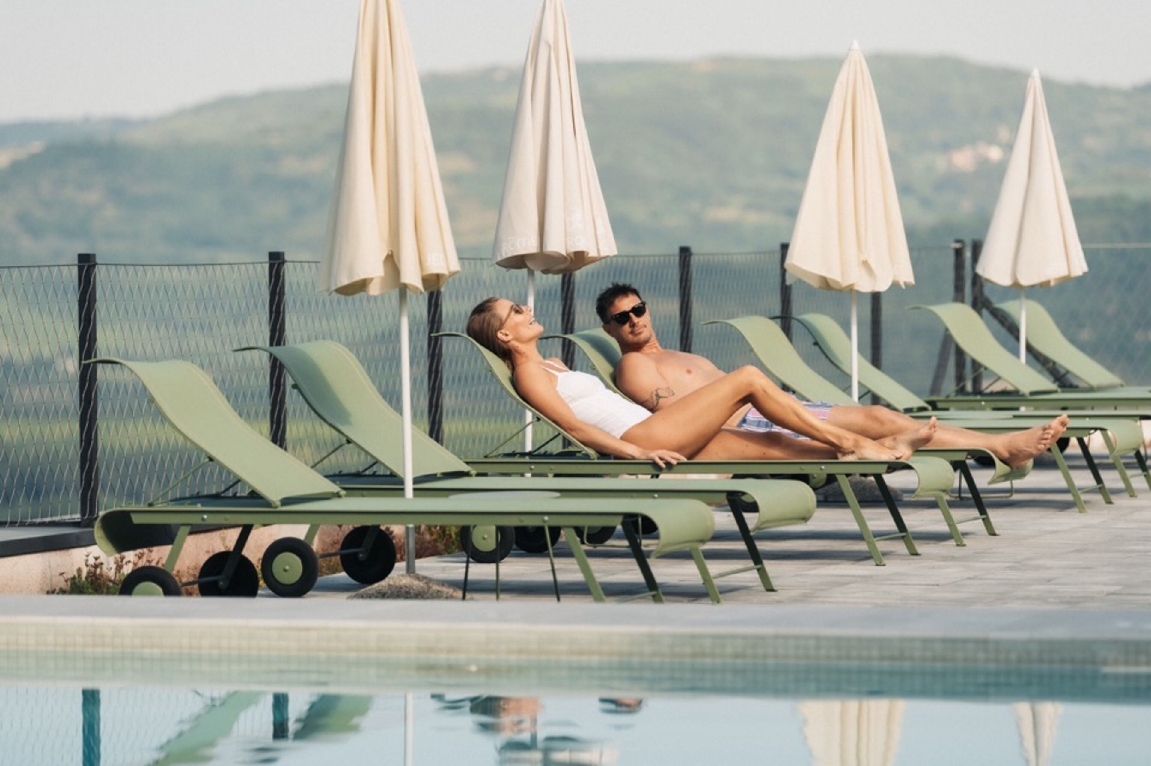 Zona junto a la piscina con sillones de descanso y sombrillas, una piscina y una valla con vistas a colinas al fondo.