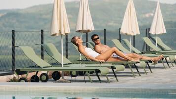 Zona junto a la piscina con sillones de descanso y sombrillas, una piscina y una valla con vistas a colinas al fondo.