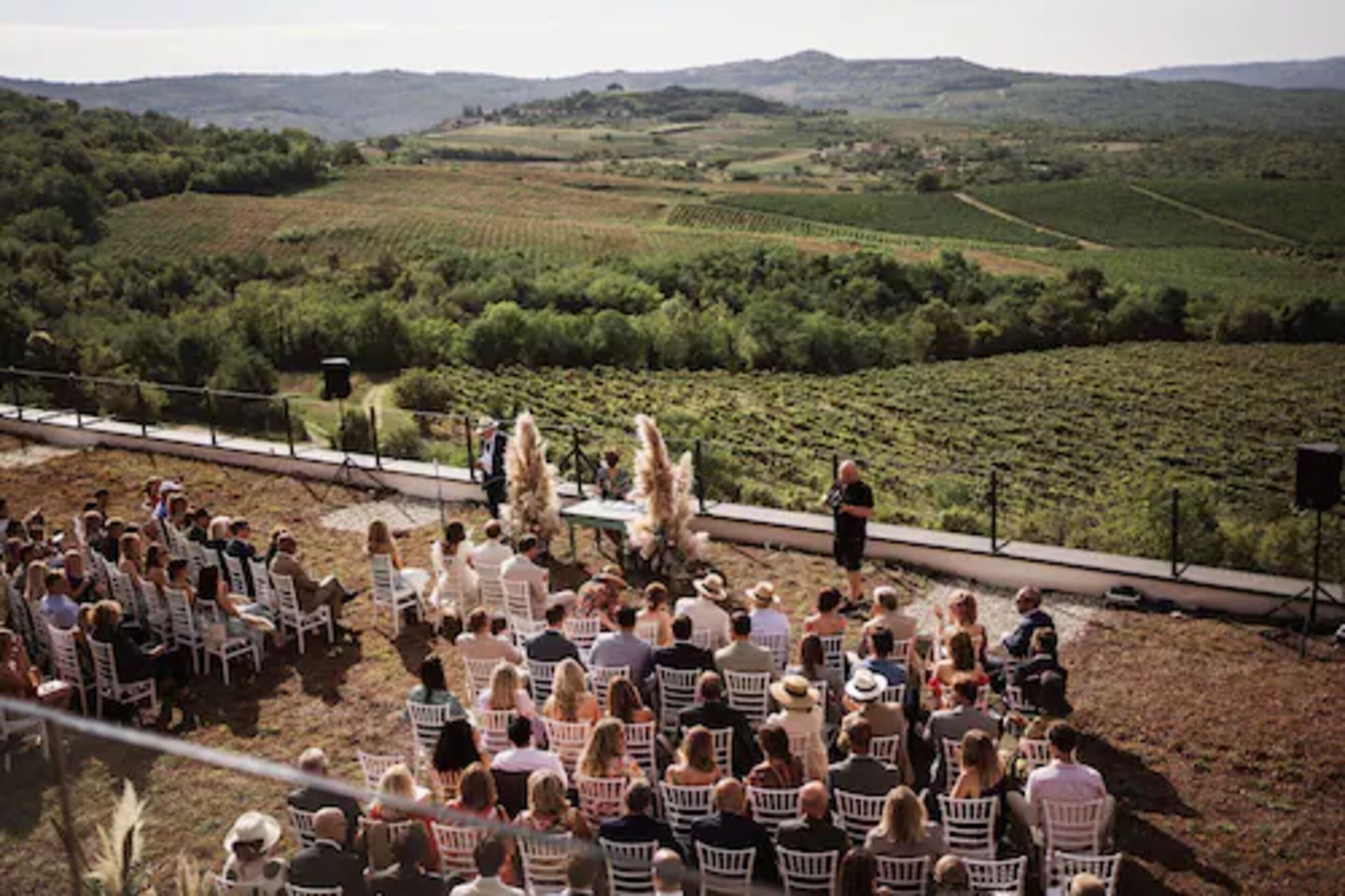 An outdoor event with attendees seated on white chairs facing a stage with a speaker.