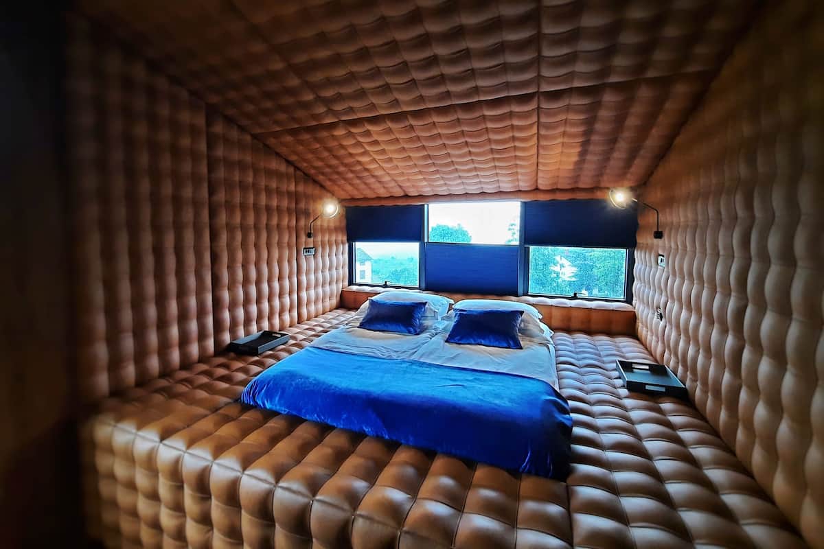 A bedroom with a tufted headboard, a bed with blue bedding, and two windows with blinds.
