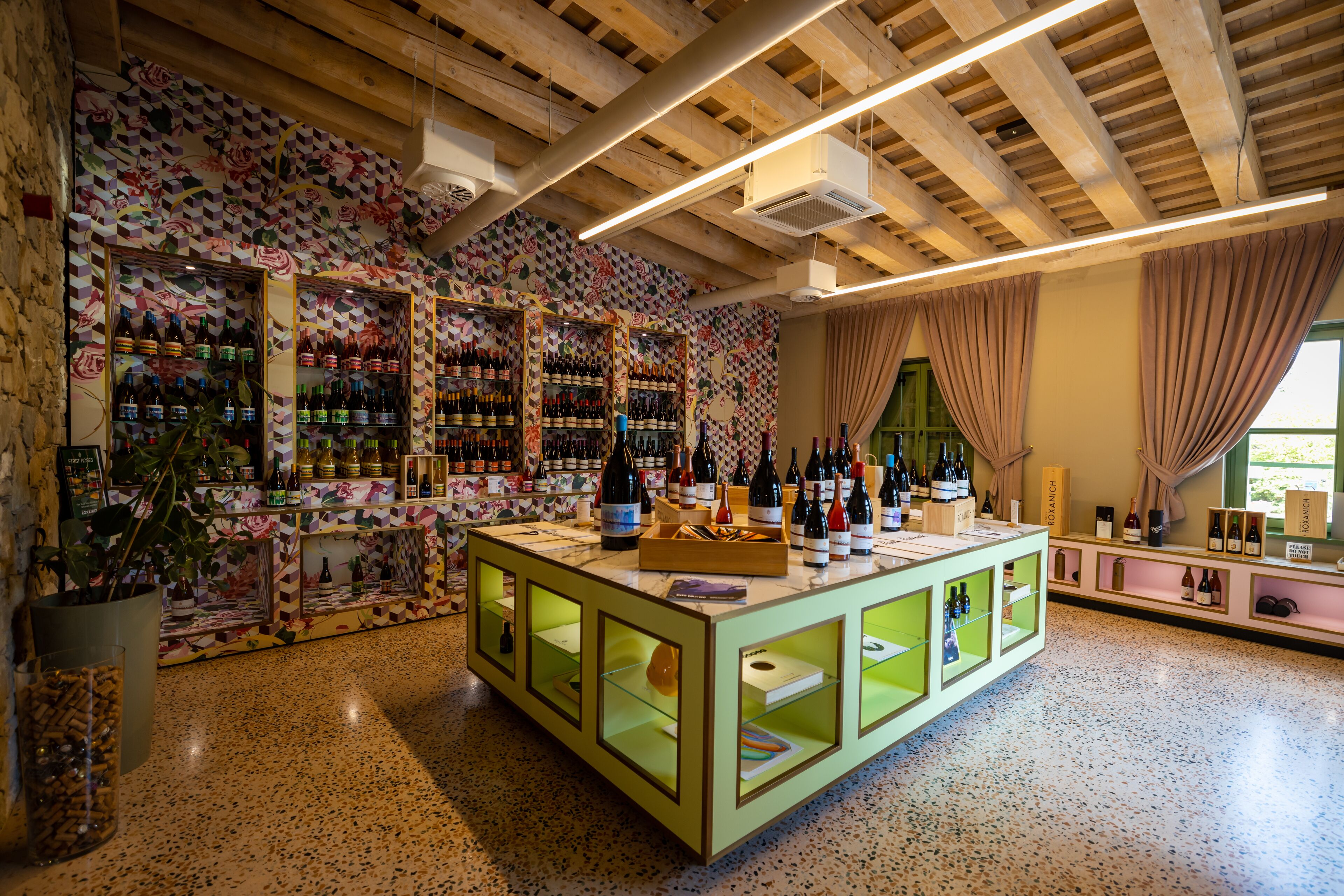 A wine shop with shelves of bottles, a central display table, and a patterned wall.