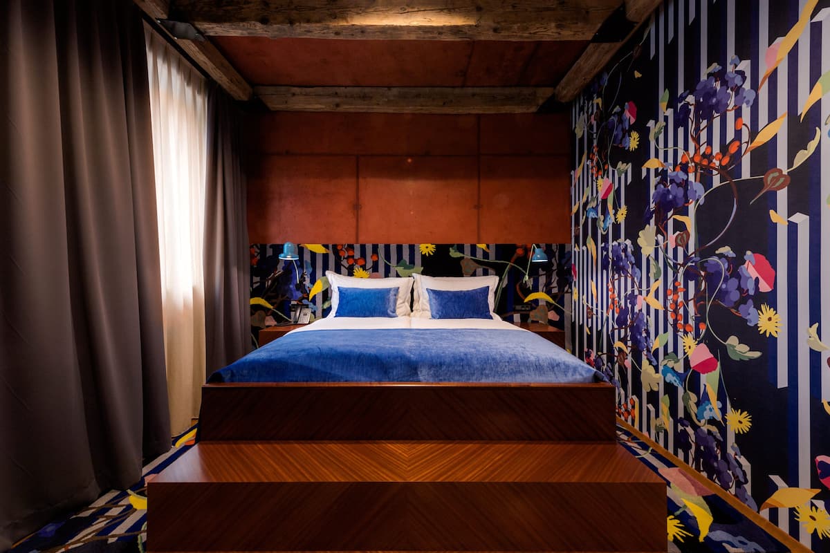 A bedroom with a bed, curtains, a wooden headboard, and a floral-patterned wall.