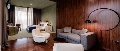 Una habitación de hotel moderna con piso de madera, un sofá gris, un sillón blanco, una mesita redonda y un televisor montado en la pared.