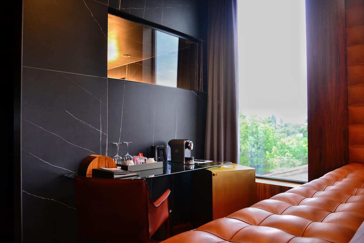 A modern hotel room with a large window, a leather sofa, a desk with a coffee maker, and a mirror.
