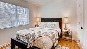 A bedroom with a bed, bedside lamps, a nightstand, a window with blinds, and a door.
