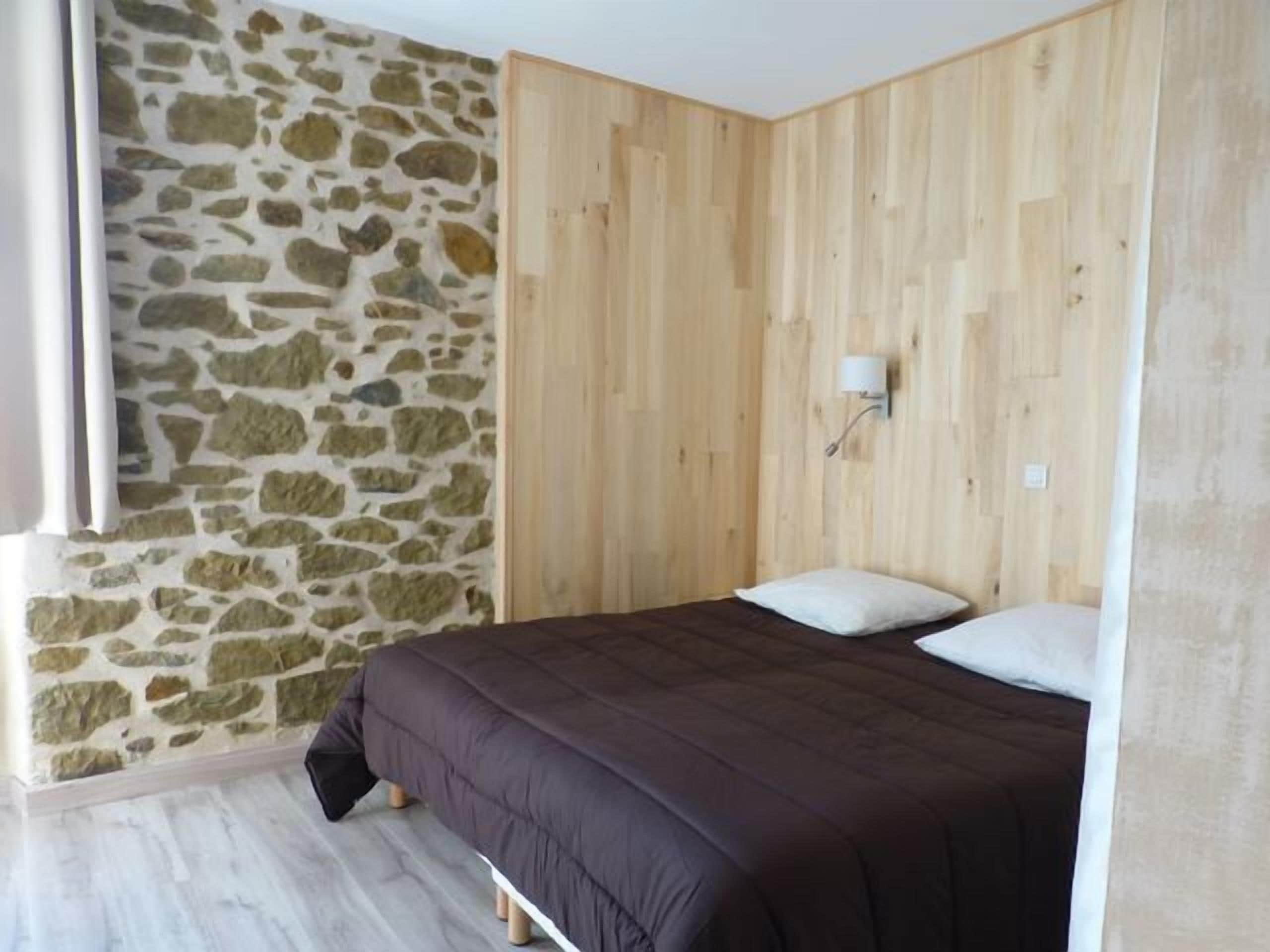 A bedroom with a stone wall, wooden paneling, a bed with a dark bedspread, and a nightstand with a lamp.
