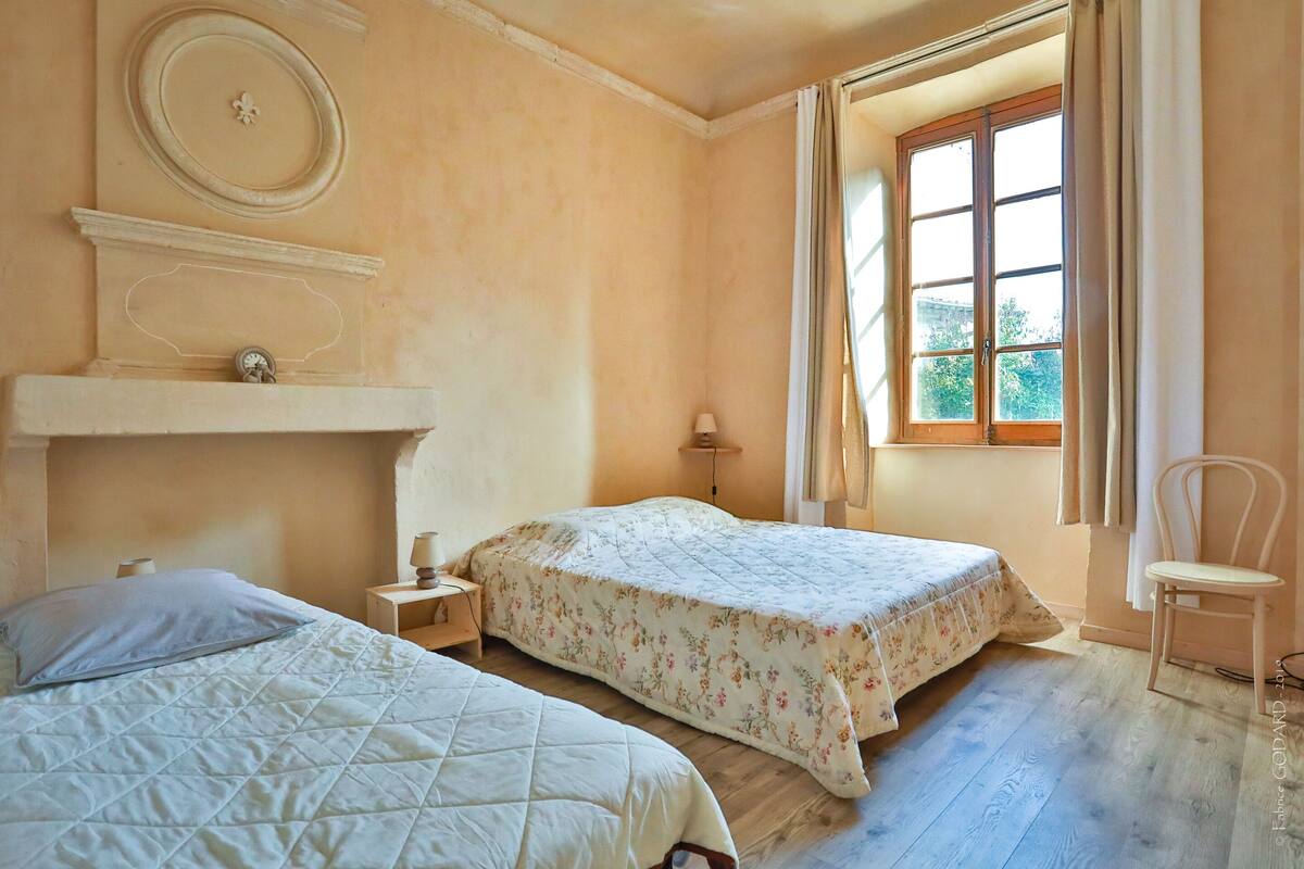 A bedroom with two beds, a window with curtains, a chair, and a small table with a lamp.