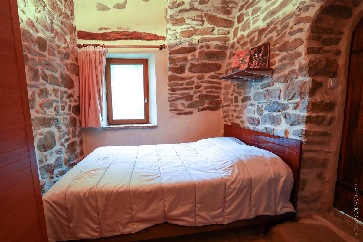 A bedroom with a stone wall, a window with a pink curtain, and a wooden bed.