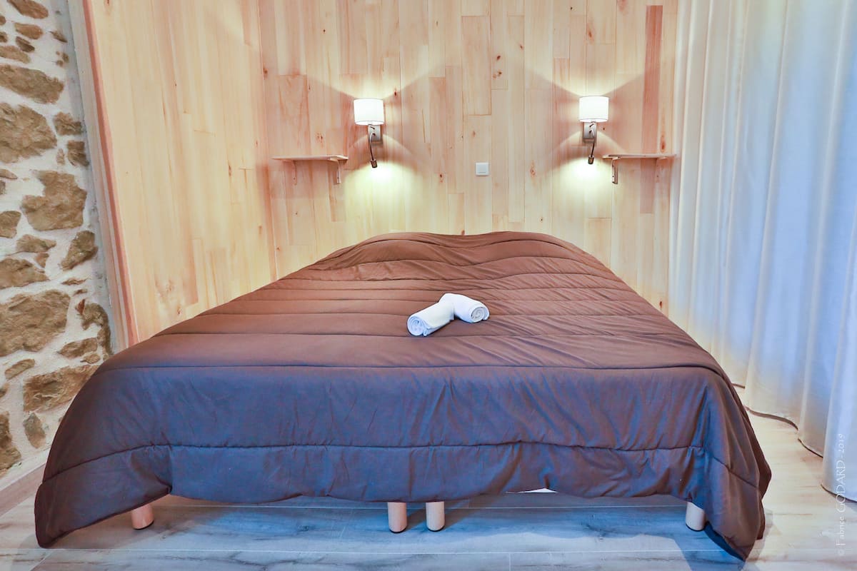 A bed with a brown bedspread and a rolled towel on it, set against a wooden wall with two wall-mounted lights.