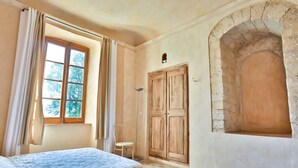 Una habitación con cama, una silla, un ventanal con cortinas y un arco de piedra.
