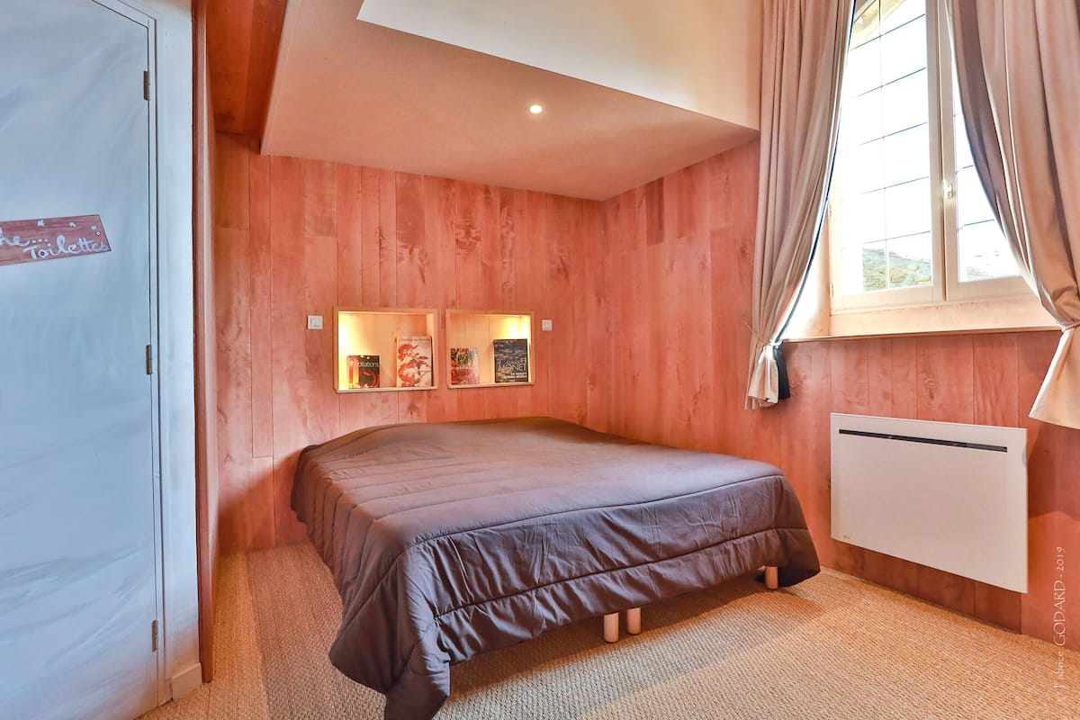 A bedroom with a bed, wooden walls, a window with curtains, and a radiator.