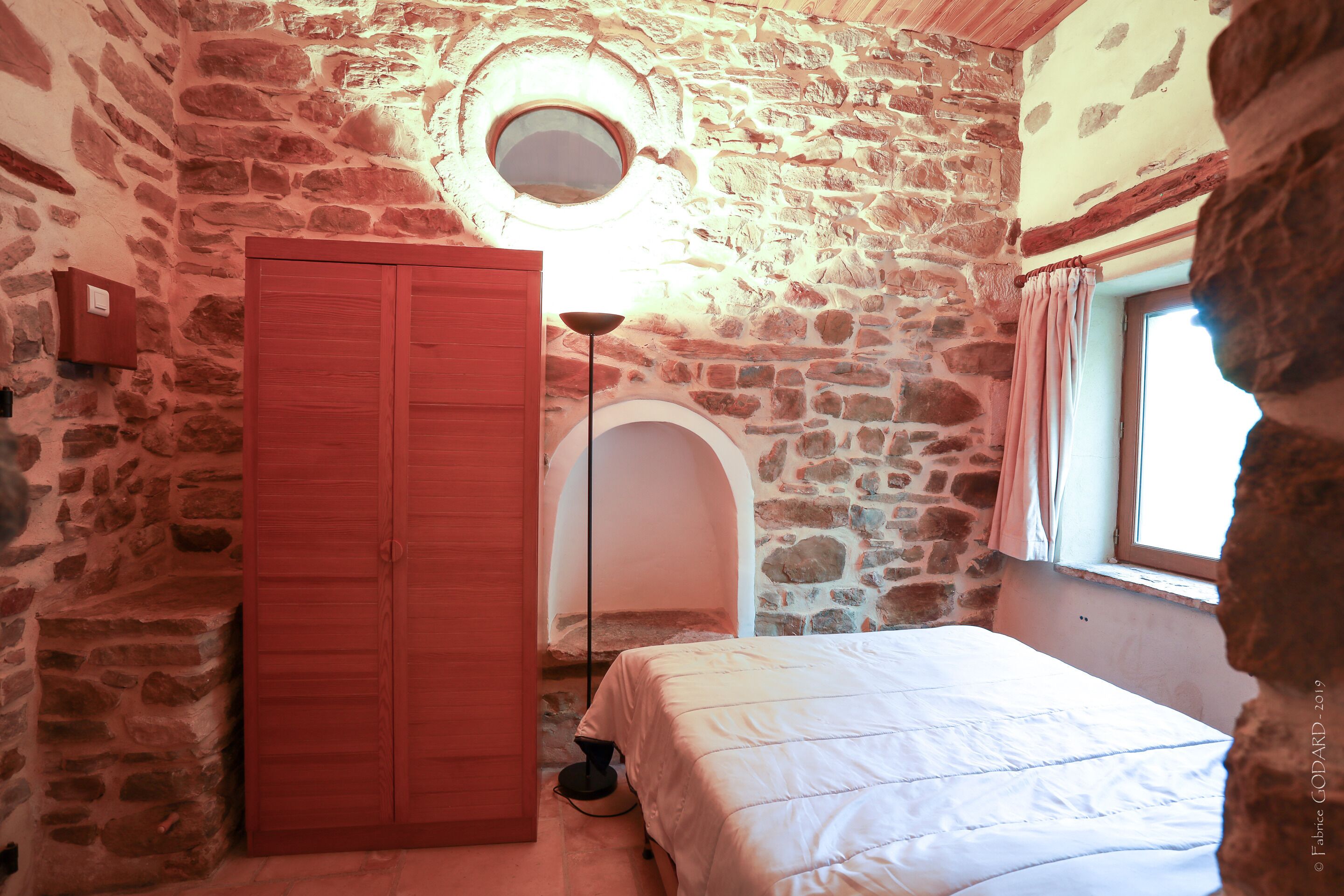 Een slaapkamer met stenen muren, een houten garderobe, een bed en een raam met gordijnen.