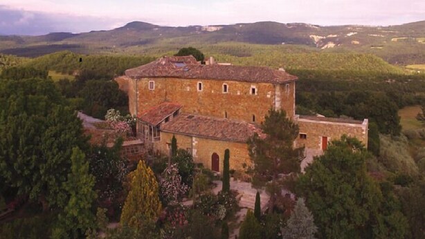 Un edificio de piedra con techo de tejas, rodeado de vegetación exuberante y colinas al fondo.