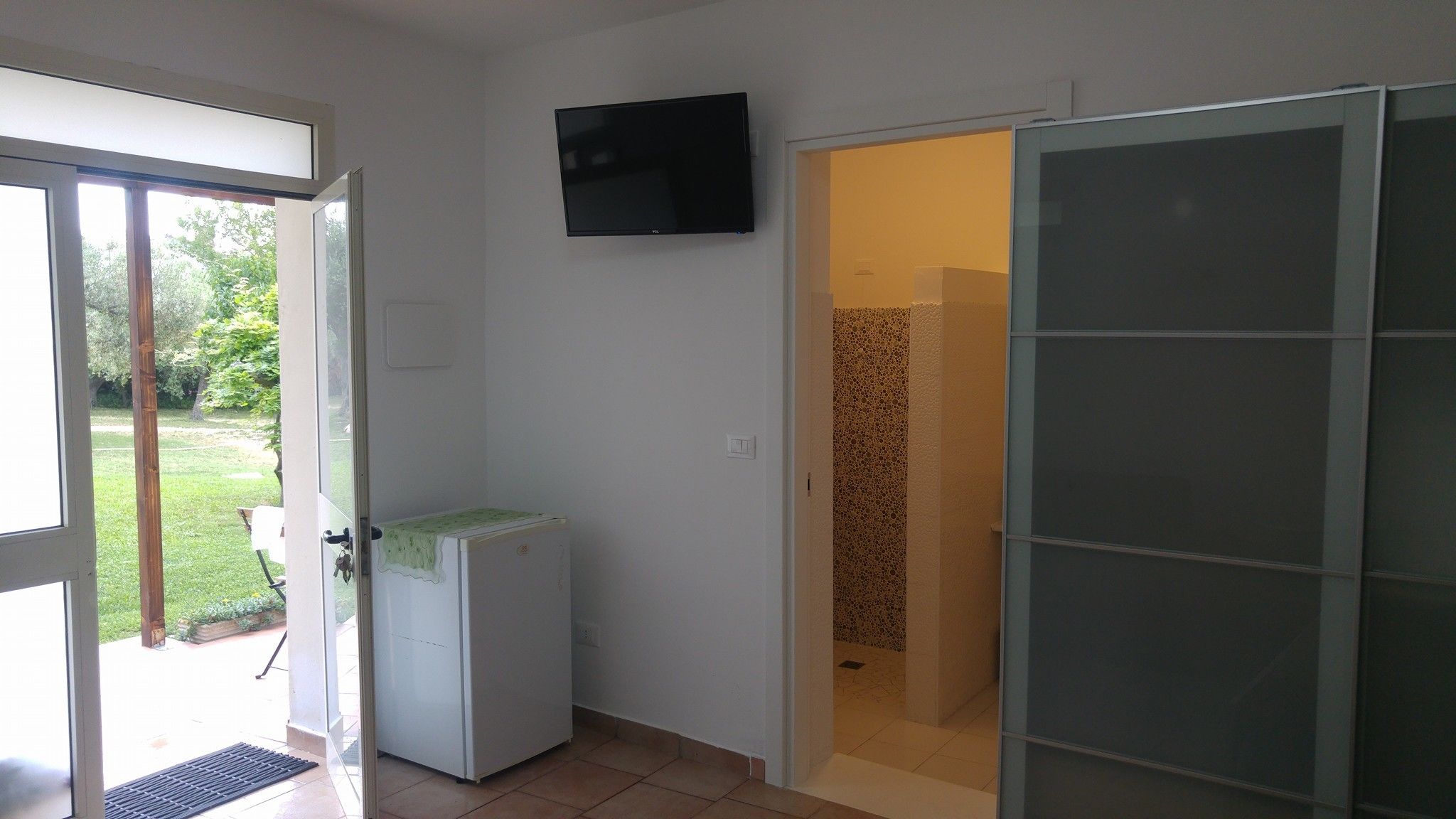 Una habitación con puerta corrediza de vidrio que da a un jardín, un refrigerador blanco pequeño, un televisor de pantalla plana montado en la pared y una puerta corrediza de vidrio hacia una zona de ducha.