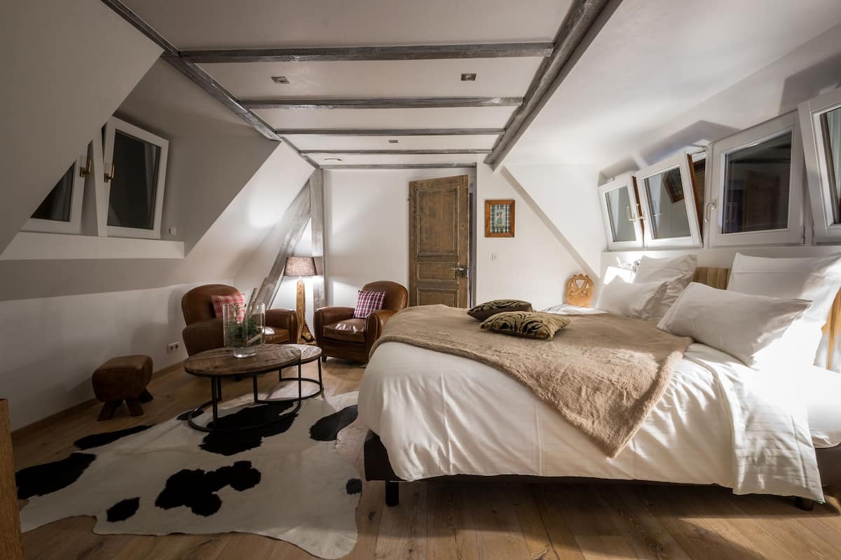 Um quarto com uma cama grande, piso de madeira, tapete de couro de vaca, uma cadeira, uma mesa e uma porta.