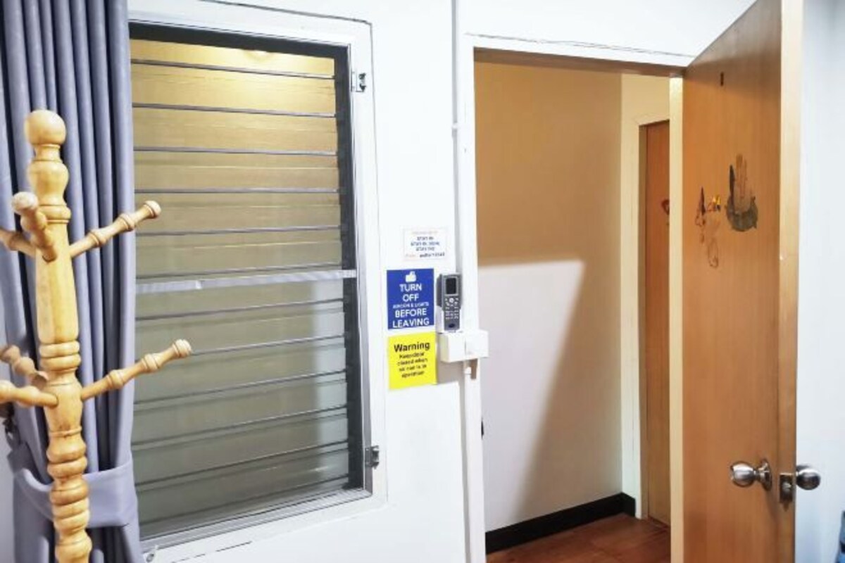 A hotel room with a window with blinds, a wooden coat rack, a door with a sign instructing to turn off the heater before leaving, and a wooden floor.