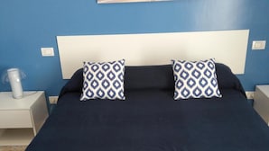 Un dormitorio con una colcha azul oscuro, dos almohadas estampadas, un cabecero blanco y un cuadro colgado en la pared que muestra una playa.