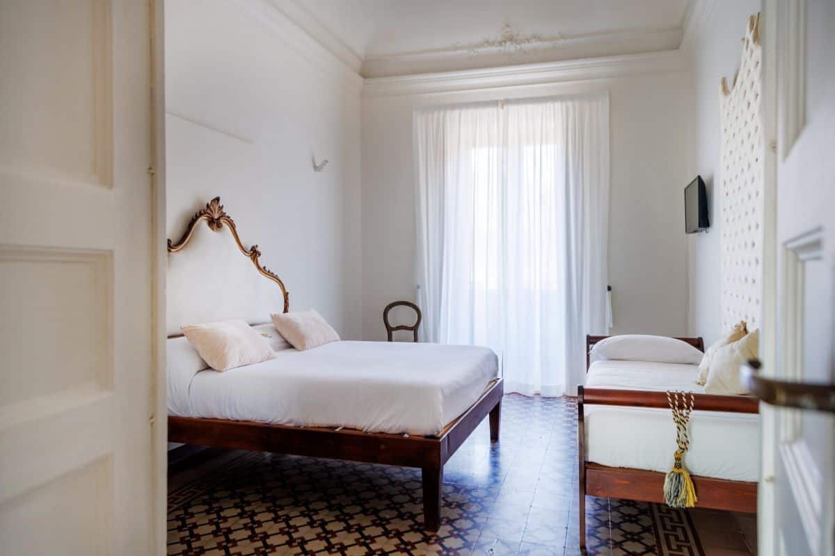A bedroom with a wooden four-poster bed, a smaller bed with a white headboard, a chair, and a patterned floor.