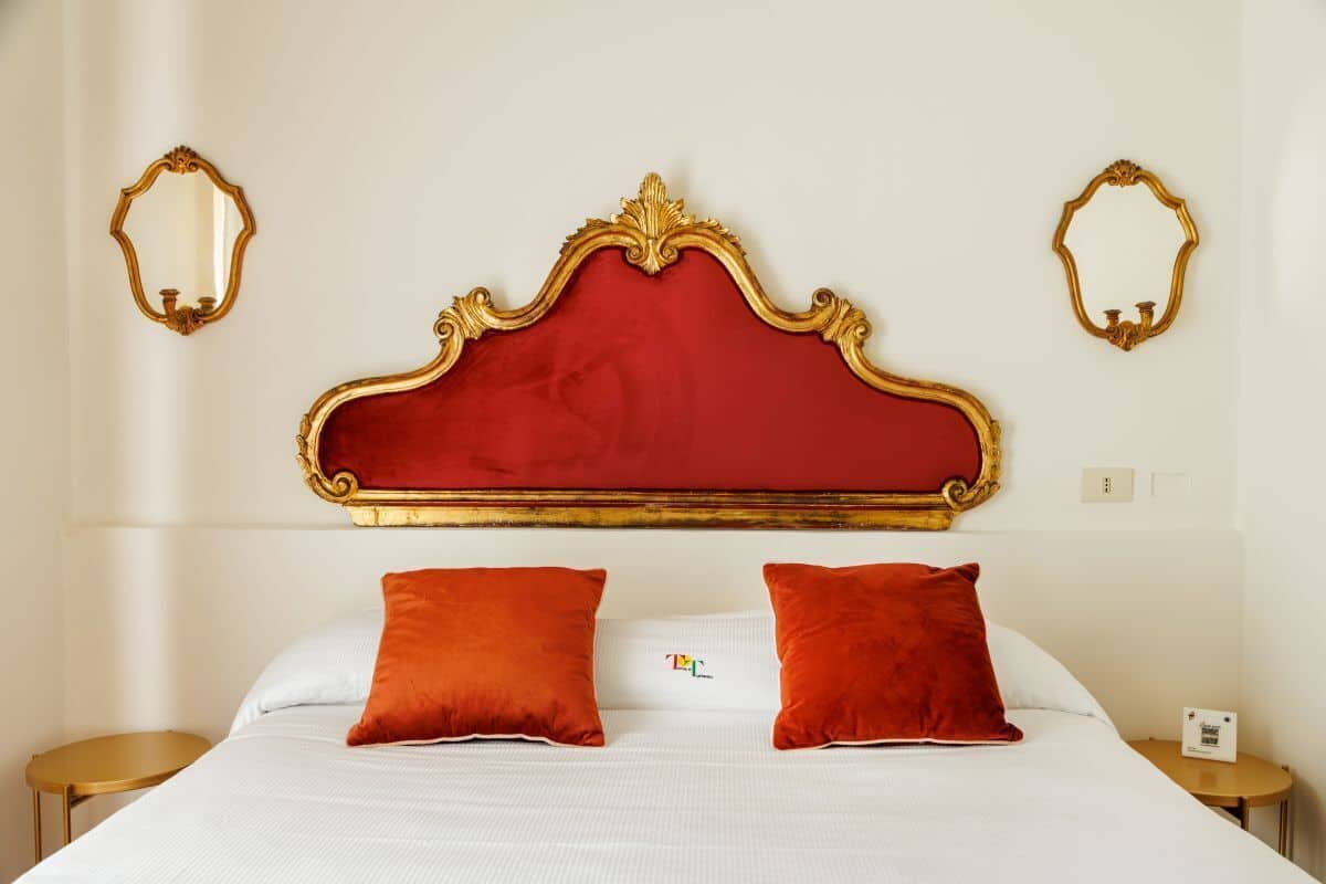 A hotel room with a large bed, two red pillows, a white bedspread, and a red upholstered headboard. There are two gold-framed mirrors on the walls.