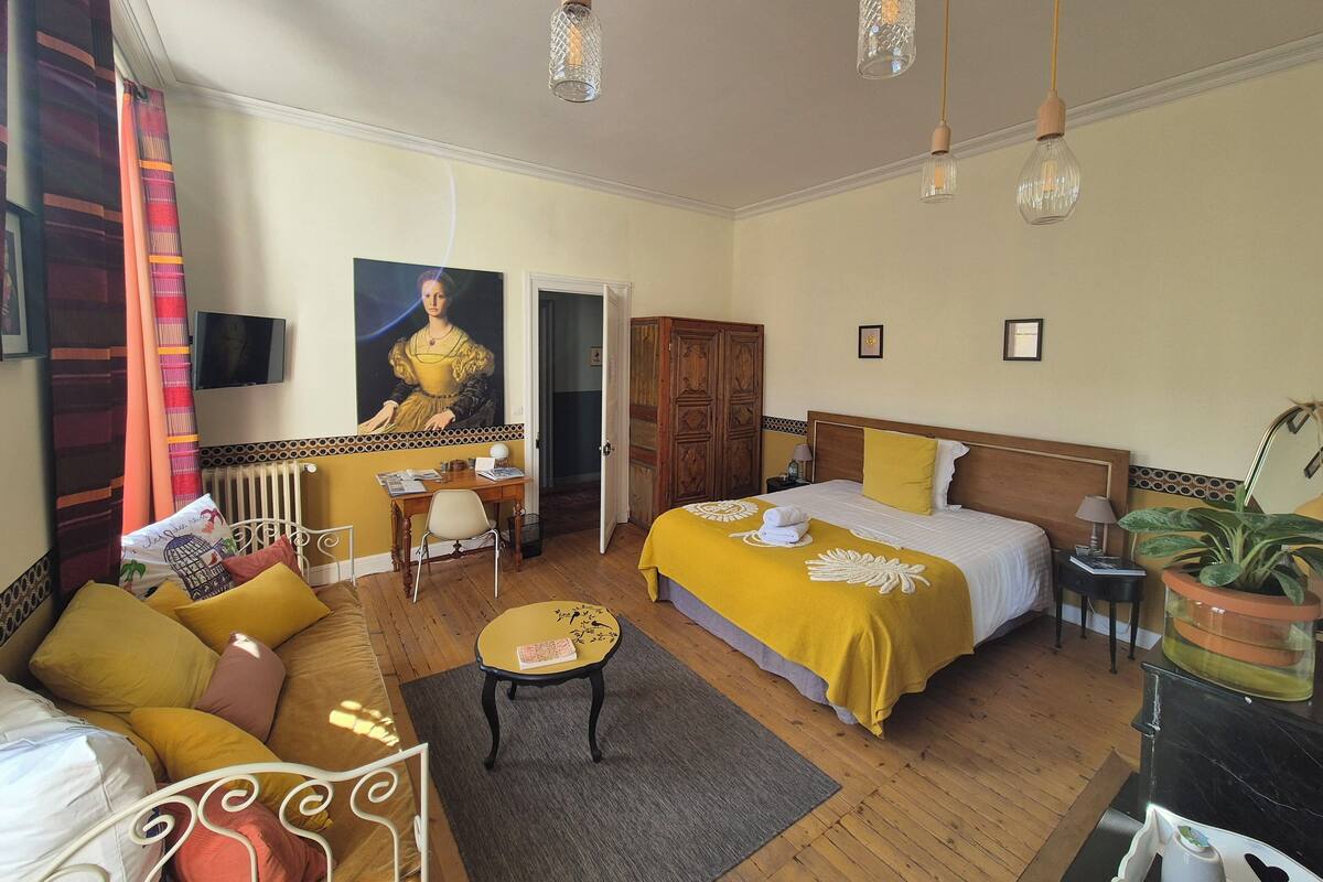 A hotel room with a bed, a sofa, a desk, and a chair. There is a painting on the wall, a potted plant, and a television.