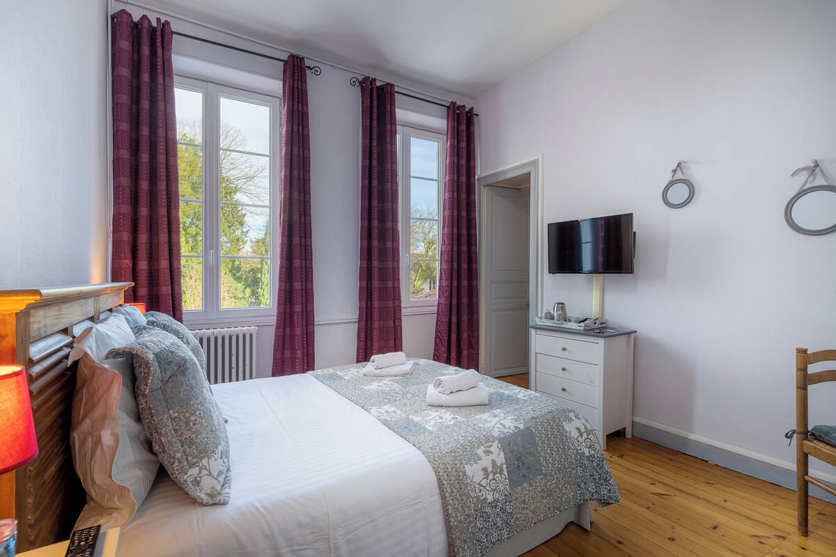 A bedroom with a bed, a bedside table with a lamp, a TV, and a mirror on the wall.