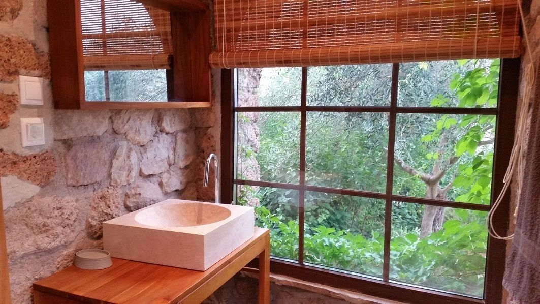 Un baño con pared de piedra, tocador de madera, un ventanal con vistas a la vegetación y una persiana de bambú.