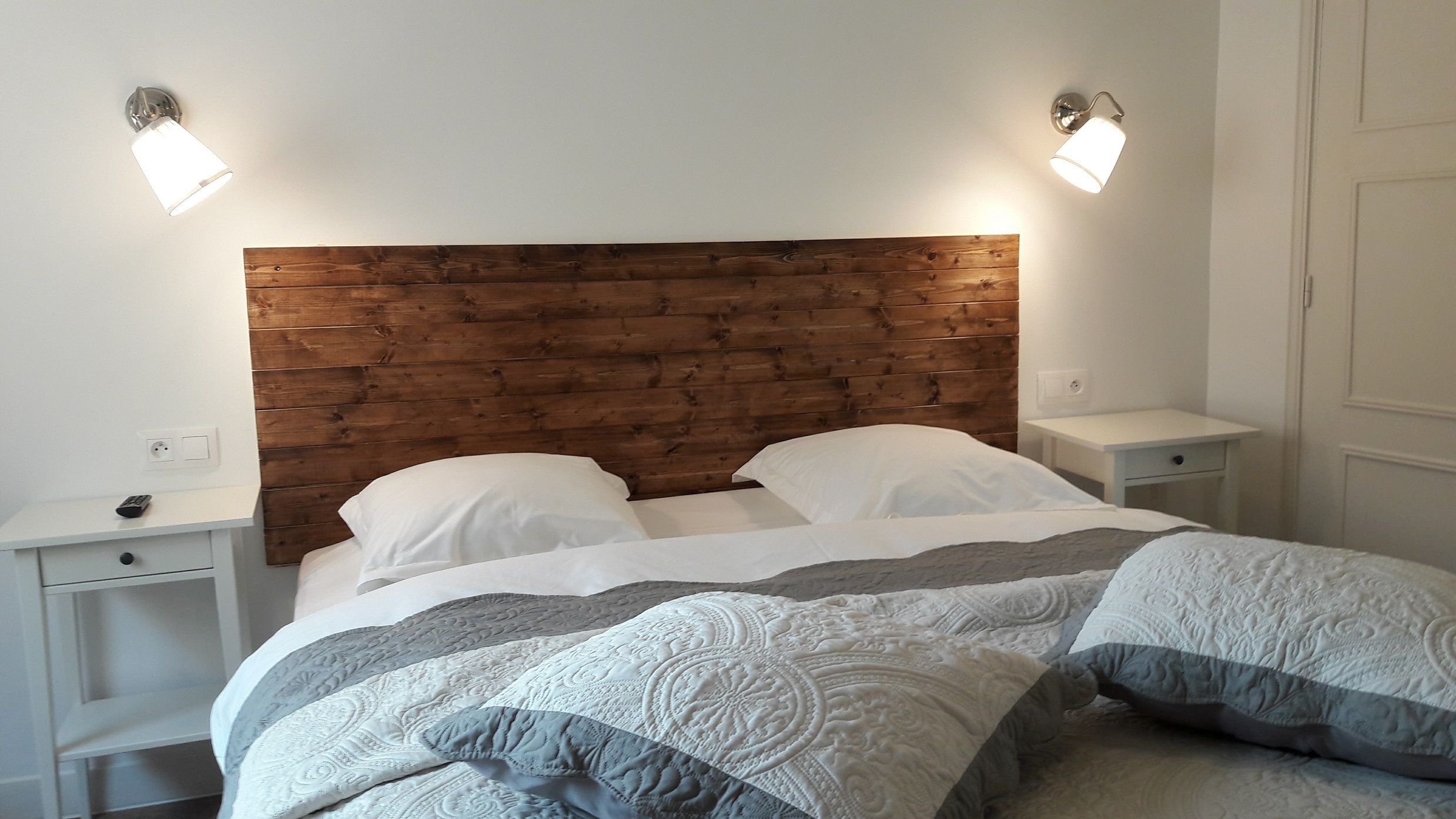 Una cama con cabecera de madera, dos mesitas de noche y lámparas fijadas a la pared.