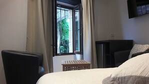 Un dormitorio con cama, una silla, un taburete de madera, una ventana con cortinas y un televisor montado en la pared.
