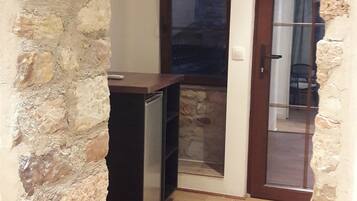 Un pasillo estrecho con una pared de piedra a un lado y suelo de madera. Una puerta da a una habitación con ventana y una silla.