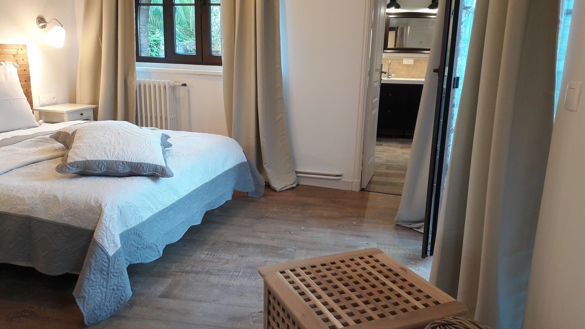 A bedroom with a bed, a nightstand, a radiator, and a view of the kitchen through an open door.