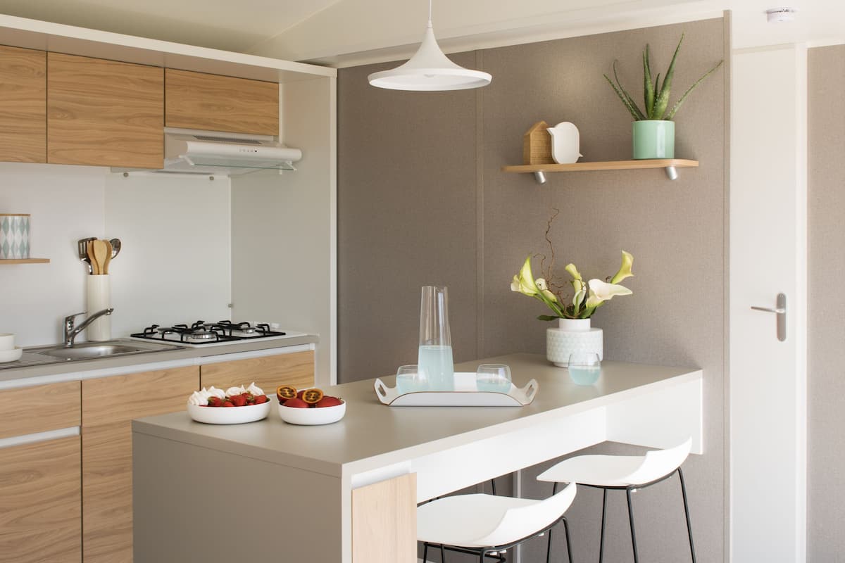 A modern kitchen with a central island, bar stools, a hanging light fixture, and a potted plant.
