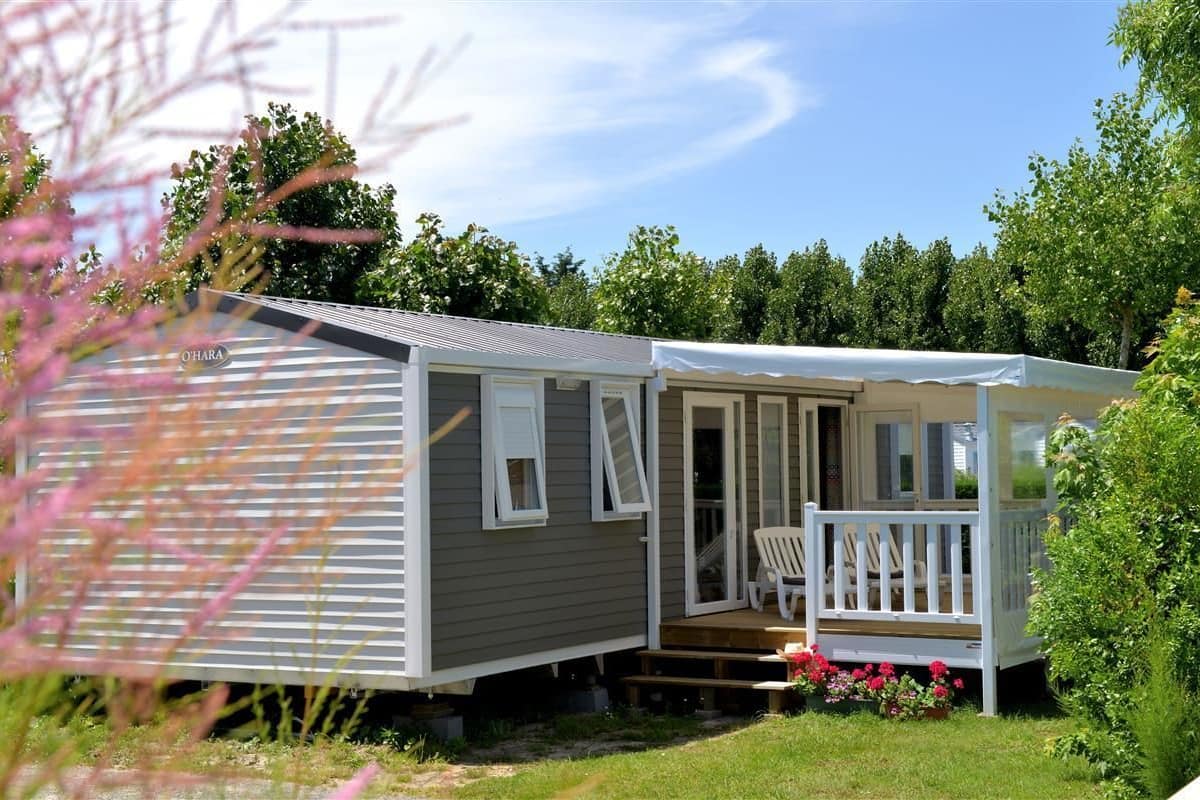 A single-story mobile home with a porch and a small deck, surrounded by greenery.