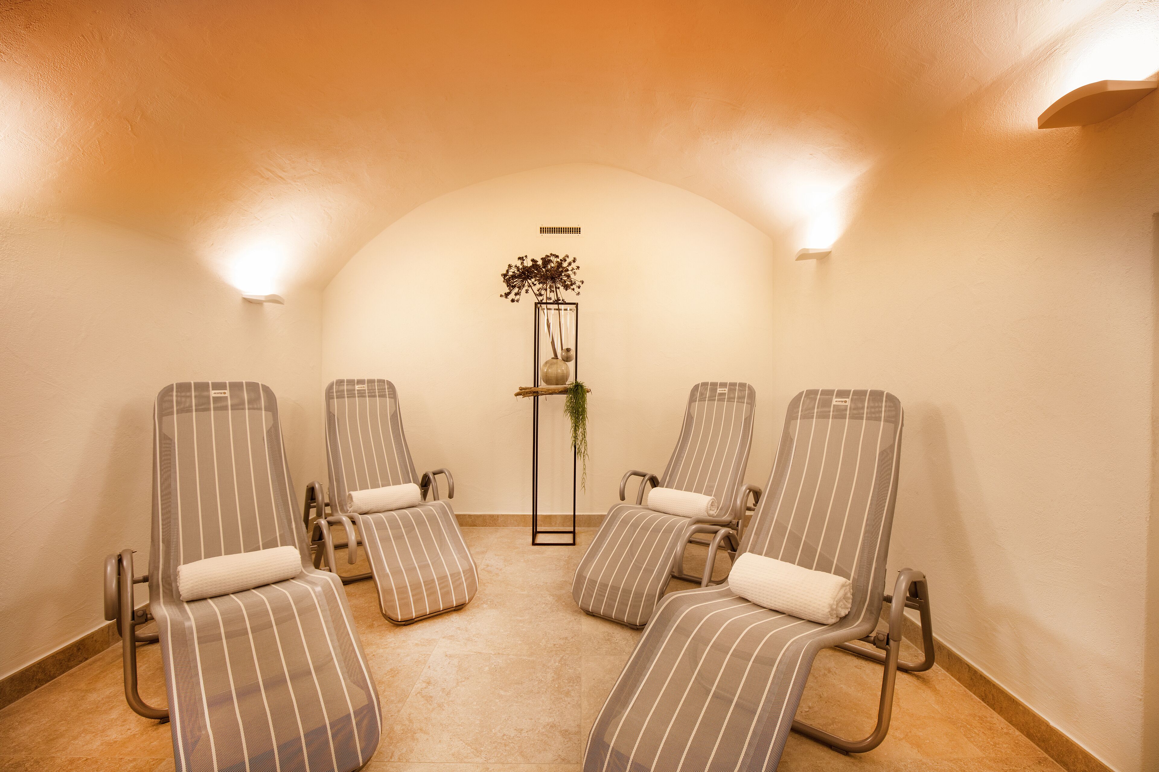 Un cuarto de spa con cuatro sillones reclinables a rayas, una planta pequeña sobre un pedestal y un suelo de baldosas.