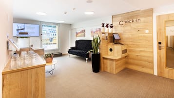 Ein Empfangsbereich mit einem Holztisch, einem schwarzen Sofa, einem 'Check-in'-Schild und einem großen Fenster.