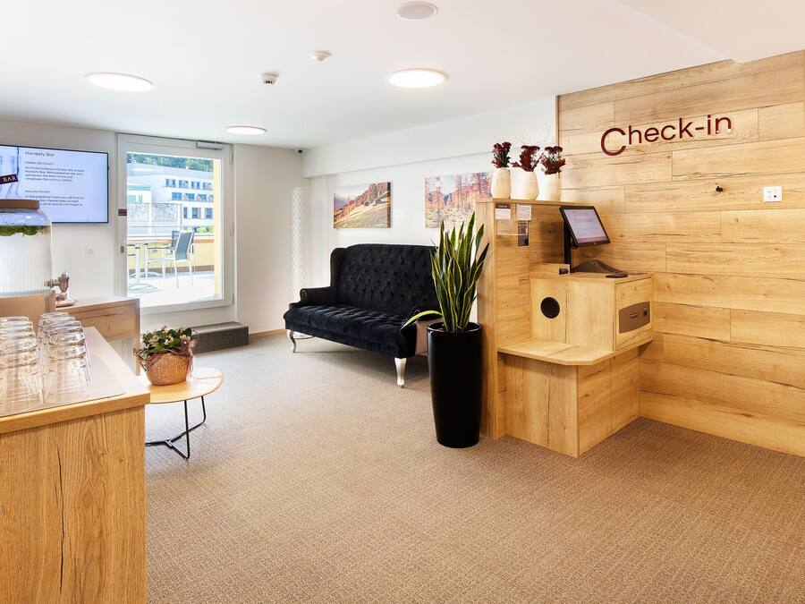 Ein Empfangsbereich mit einem Holztisch, einem schwarzen Sofa, einem 'Check-in'-Schild und einem großen Fenster.