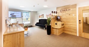 Un área de recepción con un escritorio de madera, un sofá negro, un letrero que dice 'Check-in' y un ventanal grande.